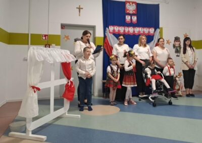 Dorośli i dzieci w sali z polskimi symbolami i flagą, niektóre w ludowych strojach.