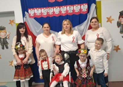 Grupa dorosłych i dzieci w strojach ludowych przy dekoracji na Święto Niepodległości Polski.