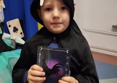 Chłopiec w czarnej pelerynie trzyma książkę z kogutem na okładce w ozdobionym pomieszczeniu.