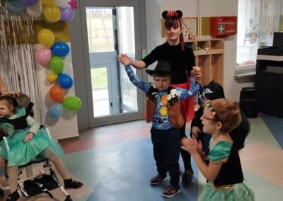 Grupa dzieci bawi się w pomieszczeniu z balonami, jedno dziecko przebrane, obok dorosły.