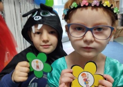 Dwoje dzieci w kostiumach z papierowymi kwiatkami, balony i dekoracje w tle.