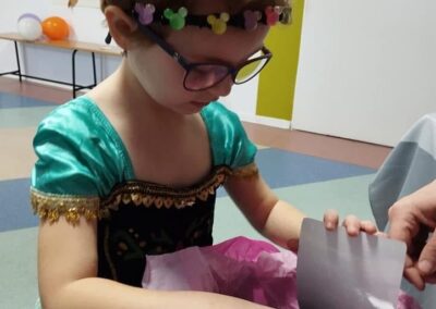 Dziewczynka w zielonej sukience, otwierająca różowy prezent w udekorowanym balonami pomieszczeniu.