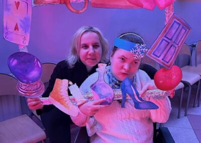 Dwie osoby z ozdobną ramką na wózku inwalidzkim, kolorowe światła, krzesła i balony w tle.