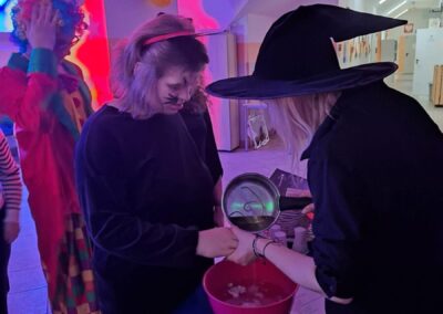 Osoby w kostiumach przy stole z miską, balony na ścianie, tematyczna impreza.