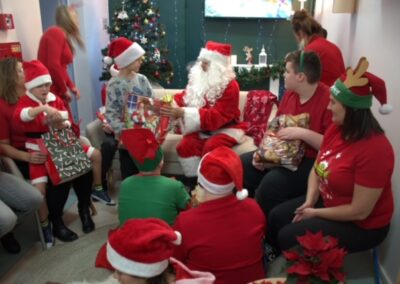 Grupa ludzi i dzieci z Mikołajem rozdającym prezenty, w tle choinka.