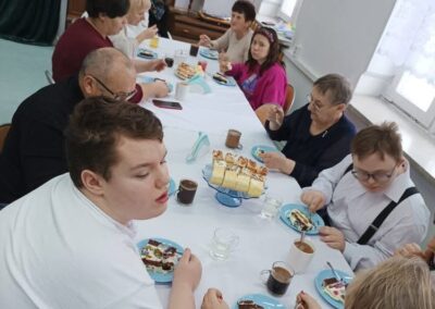 Grupa ludzi przy stole z ciastem i napojami, w jasnym pomieszczeniu z sercem na ścianie.