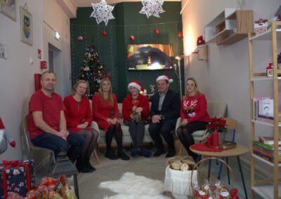 Grupa osób w czerwonych strojach w świątecznie udekorowanym pokoju z choinką i telewizorem.