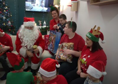 Grupa osób w świątecznych strojach z Mikołajem rozdającym prezenty w udekorowanym pokoju.