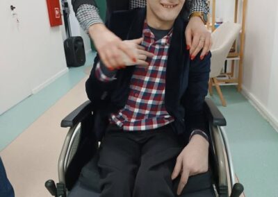 Osoba na wózku inwalidzkim w kraciastej koszuli i czapce, ktoś stoi obok w pomieszczeniu.