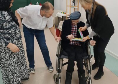 Osoby wokół starszego mężczyzny na wózku z kolorowym przedmiotem, telewizor w tle.