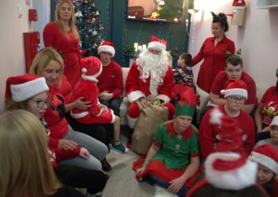 Ludzie w świątecznych ubraniach siedzą wokół Mikołaja obok udekorowanej choinki.