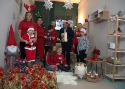 Grupa osób w świątecznych strojach w pokoju z choinką, prezentami i dekoracjami na podłodze.