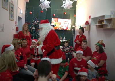 Grupa osób w strojach świątecznych w pokoju z choinką, Święty Mikołaj na pierwszym planie.