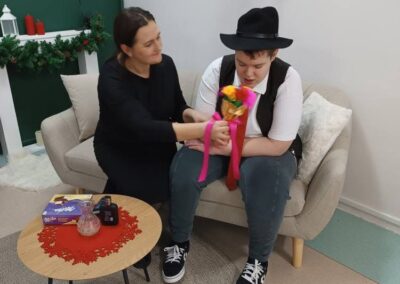 Dwie osoby na sofie, jedna trzyma lalkę, obok książka, flakon, świąteczna dekoracja w tle.