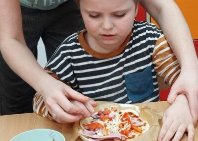 Chłopiec przy stole z pizzą, kobieta pomaga układać składniki.