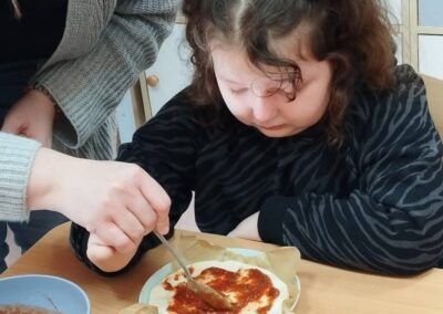 Dziecko z pomocą dorosłego nakłada sos na ciasto przy stole, obok miski z dodatkami.