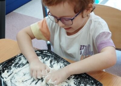 Dziecko w okularach bawi się mąką na czarnej tacy, w tle przedmioty kuchenne.