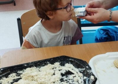 Dziecko w okularach siedzi przy stole z ciastem i wącha coś trzymanego przez dorosłego.