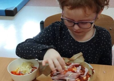 Dziecko w okularach przygotowuje pizzę przy stole z miseczką warzyw i sera obok.