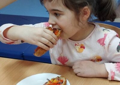 Dziewczynka je kanapkę przy drewnianym stole; talerz na białym talerzu, kolorowa bluzka.