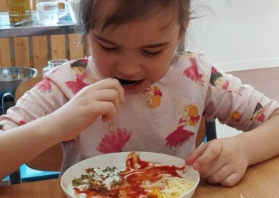 Dziewczynka je makaron z sosem pomidorowym przy drewnianym stole, w tle kuchnia.