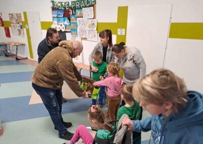Dorośli i dzieci w klasie z pracami plastycznymi, ktoś trzyma pudełko z czekoladkami.
