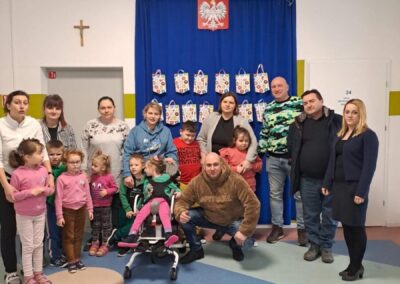 Grupa osób w sali z niebieską zasłoną, torbami na prezenty i godłem Polski na ścianie.