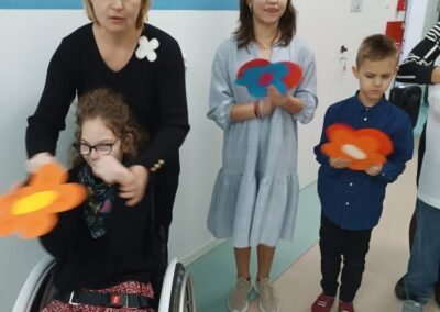 Osoby na korytarzu trzymające papierowe kwiaty, jedna na wózku inwalidzkim.