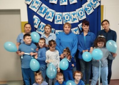 Grupa w niebieskim z balonami, dekoracja Dnia Świadomości Autyzmu, godło Polski w tle.