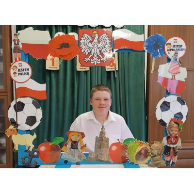Chłopiec siedzi przed ramą ozdobioną symbolami Polski, flagą, godłem, piłkami, postaciami i Pałacem Kultury.
