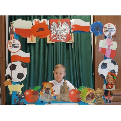 Chłopiec za ramą ozdobioną symbolami Polski, flagą, godłem, piłkami i ludowymi figurkami.