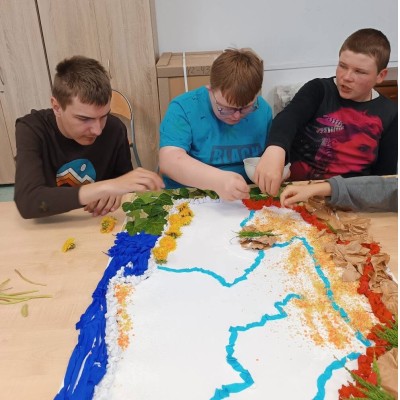 Trzech chłopców tworzy mapę z liści, kwiatów, papieru i innych materiałów przy stole.