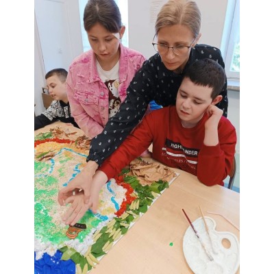 Dzieci i dorosła osoba tworzą projekt plastyczny przy stole z wykorzystaniem liści i innych materiałów.