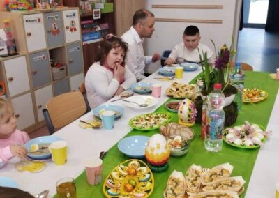 Grupa dzieci i dorosły przy stole z potrawami, świątecznymi jajkami, zabawki w tle.
