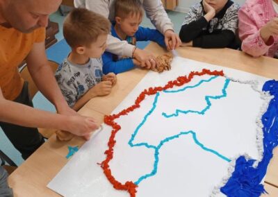 Dzieci i dorośli tworzą model mapy z kolorowych materiałów na stole w sali.