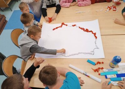 Dzieci układają obrys mapy Polski z czerwonych i białych papierowych kulek na stole.