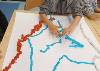 Dzieci tworzą kontury mapy z czerwonych, białych i niebieskich kawałków papieru na stole.