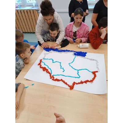 Dzieci wraz z opiekunem dekorują mapę Polski kolorową bibułą przy stole.