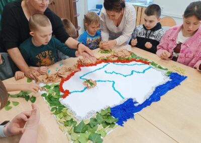 Dzieci i opiekunowie układają mapę z kolorowych materiałów i liści na stole w pomieszczeniu.