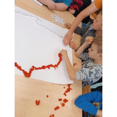 Dzieci układają czerwone i białe kulki papieru na konturze mapy Polski na stole.