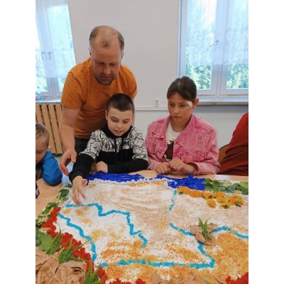 Dzieci z nauczycielem przy stole z kolorową mapą wykonaną z liści, kwiatów i ziarna.