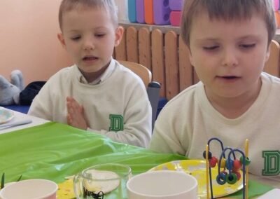 Dwaj chłopcy przy stole z zielonym obrusem, kubkami i zabawką, w tle duże klocki.