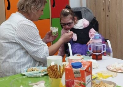 Dorosły karmi dziecko na wózku, na stole przekąski, sok, kolorowe szafki w tle.