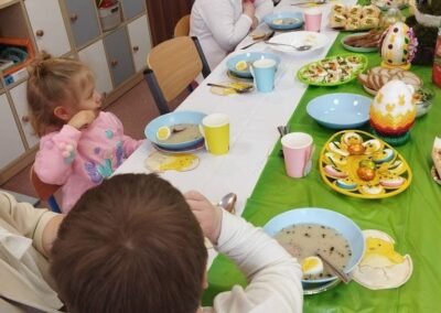 Dzieci i dorośli jedzą przy udekorowanym stole z wielkanocnymi ozdobami w przedszkolu.