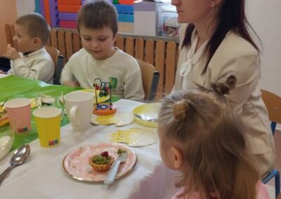 Grupa dzieci i dorosły przy stole z talerzykami i kubkami, w tle zabawki i okno.