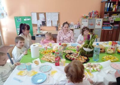 Dzieci i dorośli przy stole z jedzeniem w jasnym pokoju z meblami i tablicą informacyjną.