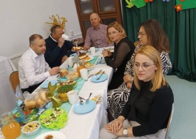 Grupa osób przy stole z potrawami i wielkanocnymi dekoracjami na zielonym tle.