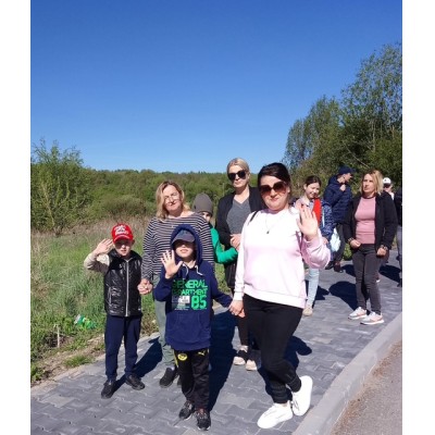 Grupa dorosłych i dzieci idzie chodnikiem obok zielonego terenu pod słonecznym niebem.