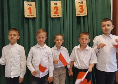 Piątka chłopców w białych koszulach z flagami Polski na tle dekoracji Święta Majowe.
