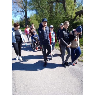 Grupa osób idzie chodnikiem, wśród nich jedna osoba na wózku inwalidzkim, w słoneczny dzień.